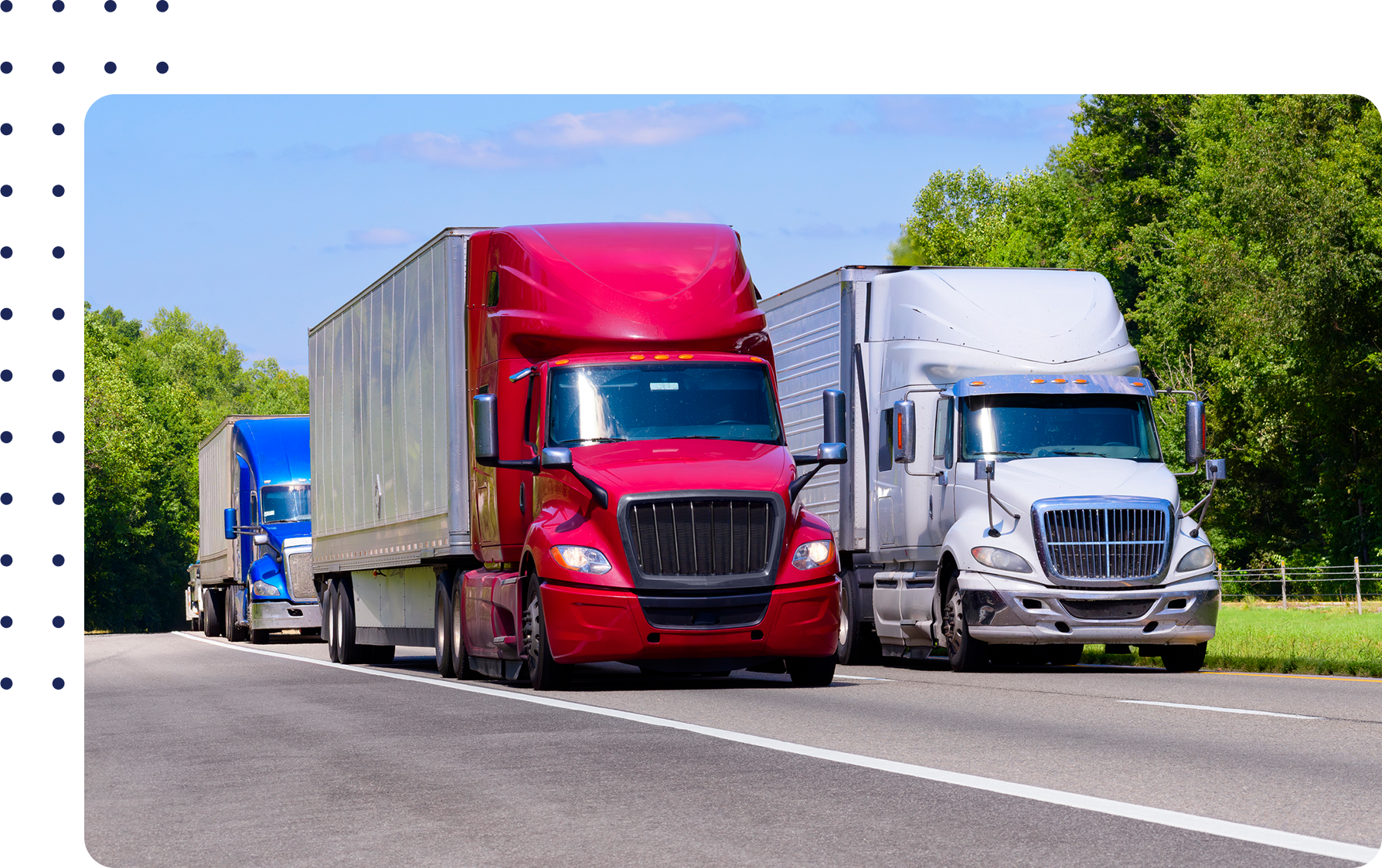Three semi-trucks, colored blue, red, and silver, drive on a highway surrounded by green trees under a clear blue sky.